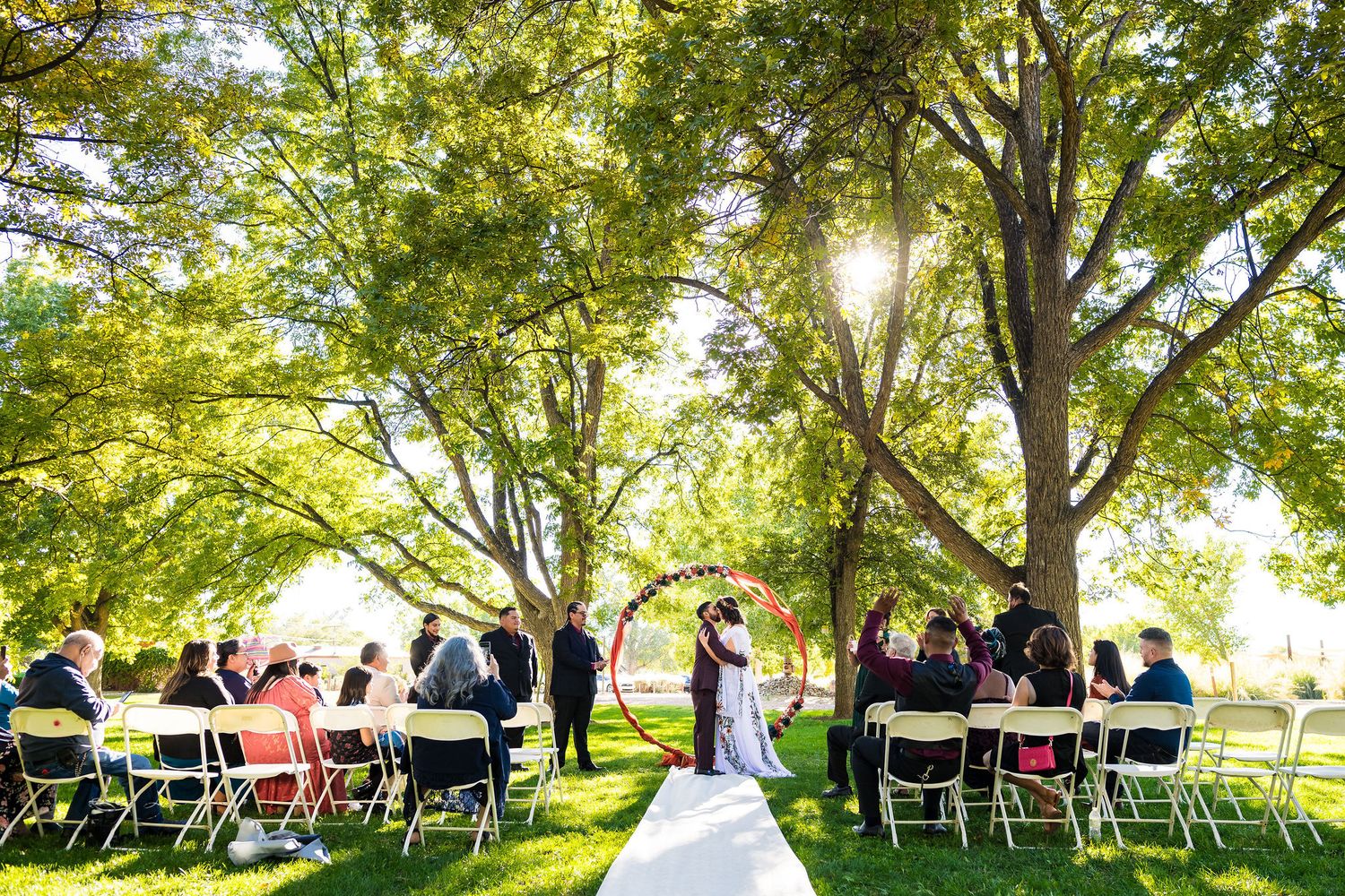 Outdoor wedding ceremony under large green trees with guests seated on white chairs in Albuquerque, New Mexico.