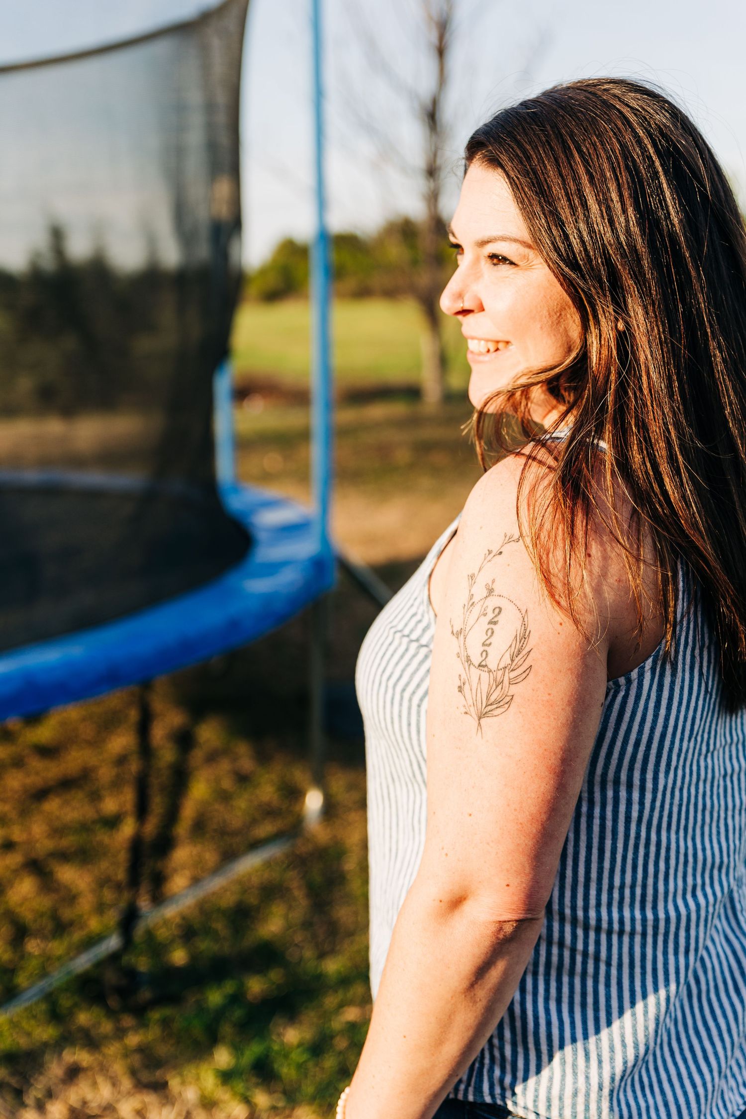 Smiling brunette woman with shoulder tattoo in striped top standing outdoors near blue trampoline in golden sunlight.
