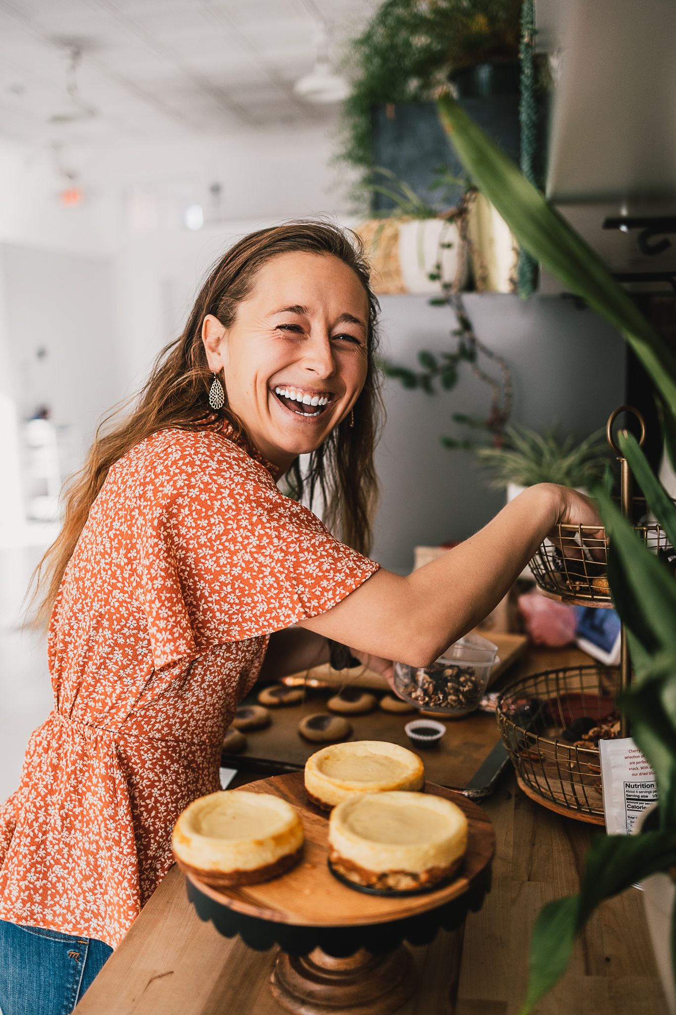 Laughing woman in floral top arranges baked goods on counter in bright plant-filled cafe.