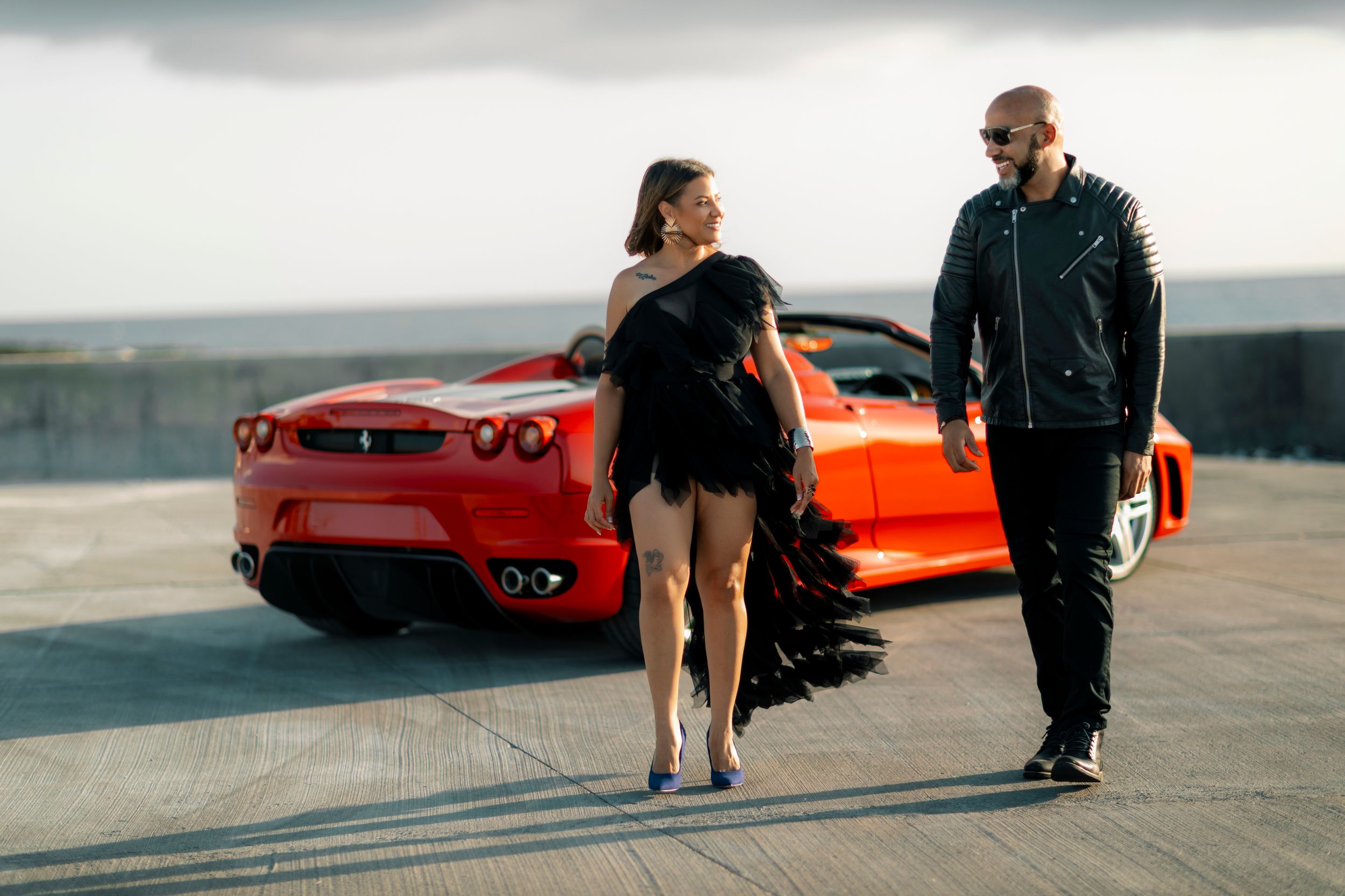 A couple poses in front of a red Ferrari sports car on an overcast day.