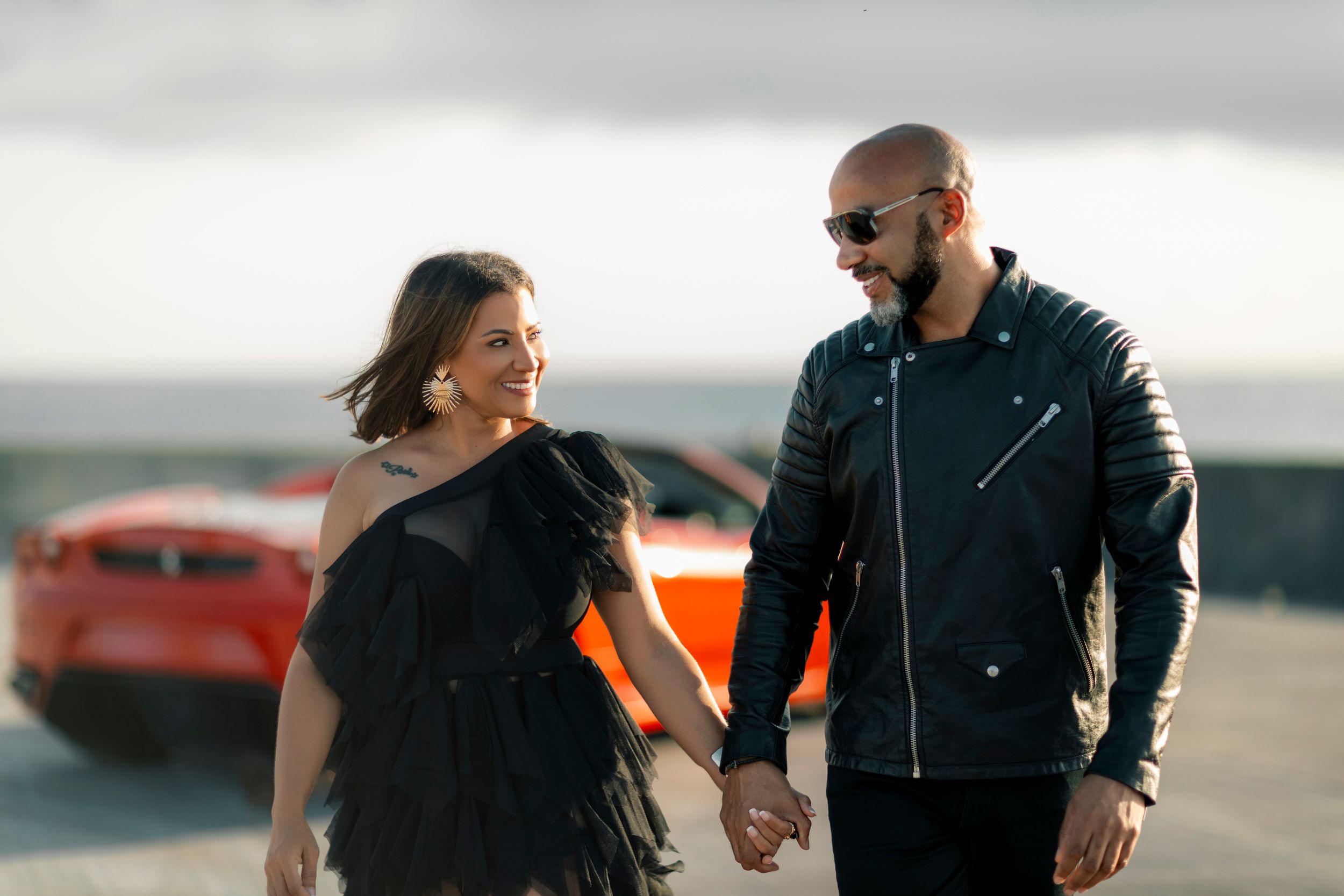 A couple in black outfits holding hands with a red sports car in the background.