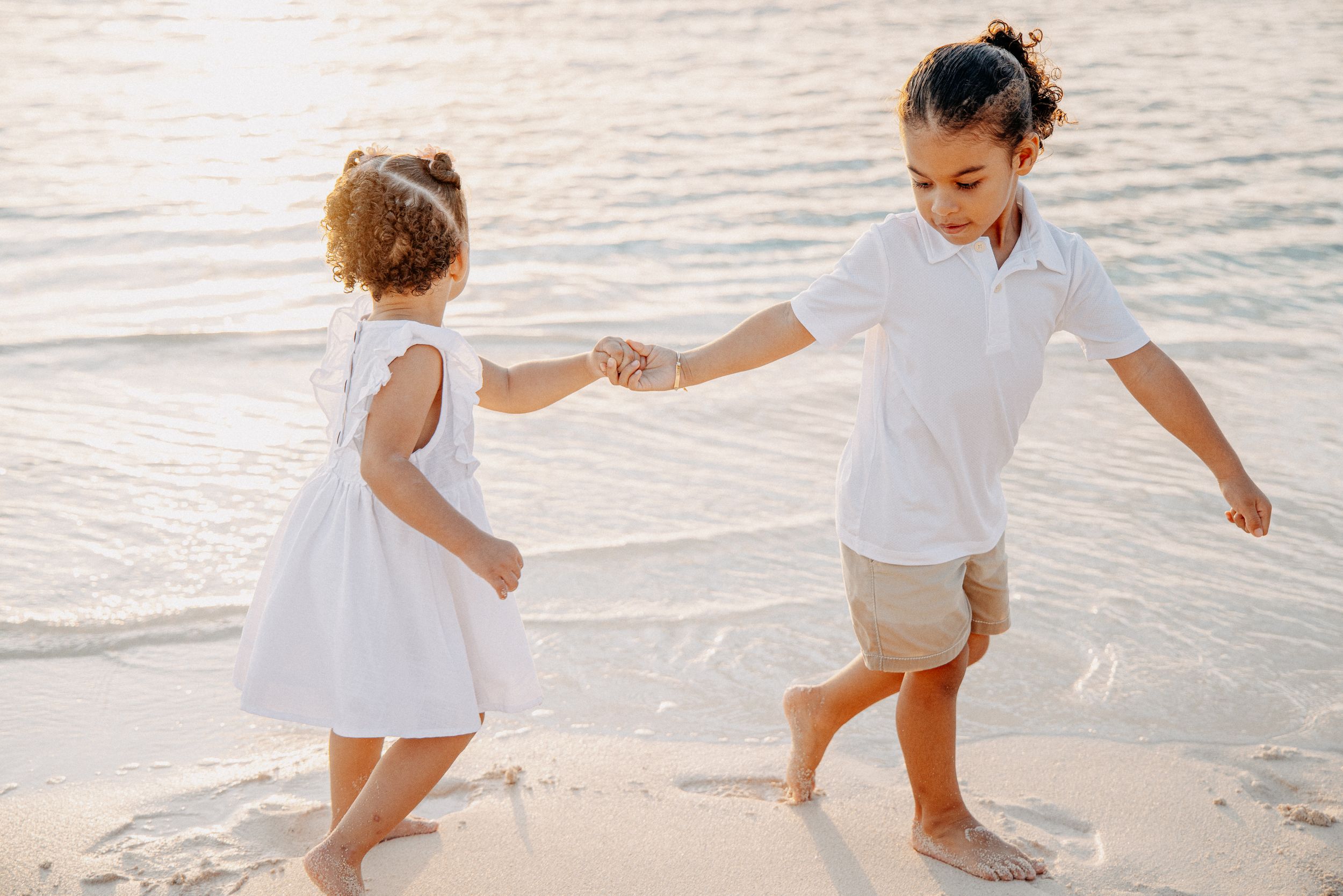 Two people in white outfits playing together at the edge of the ocean on a sunny beach.