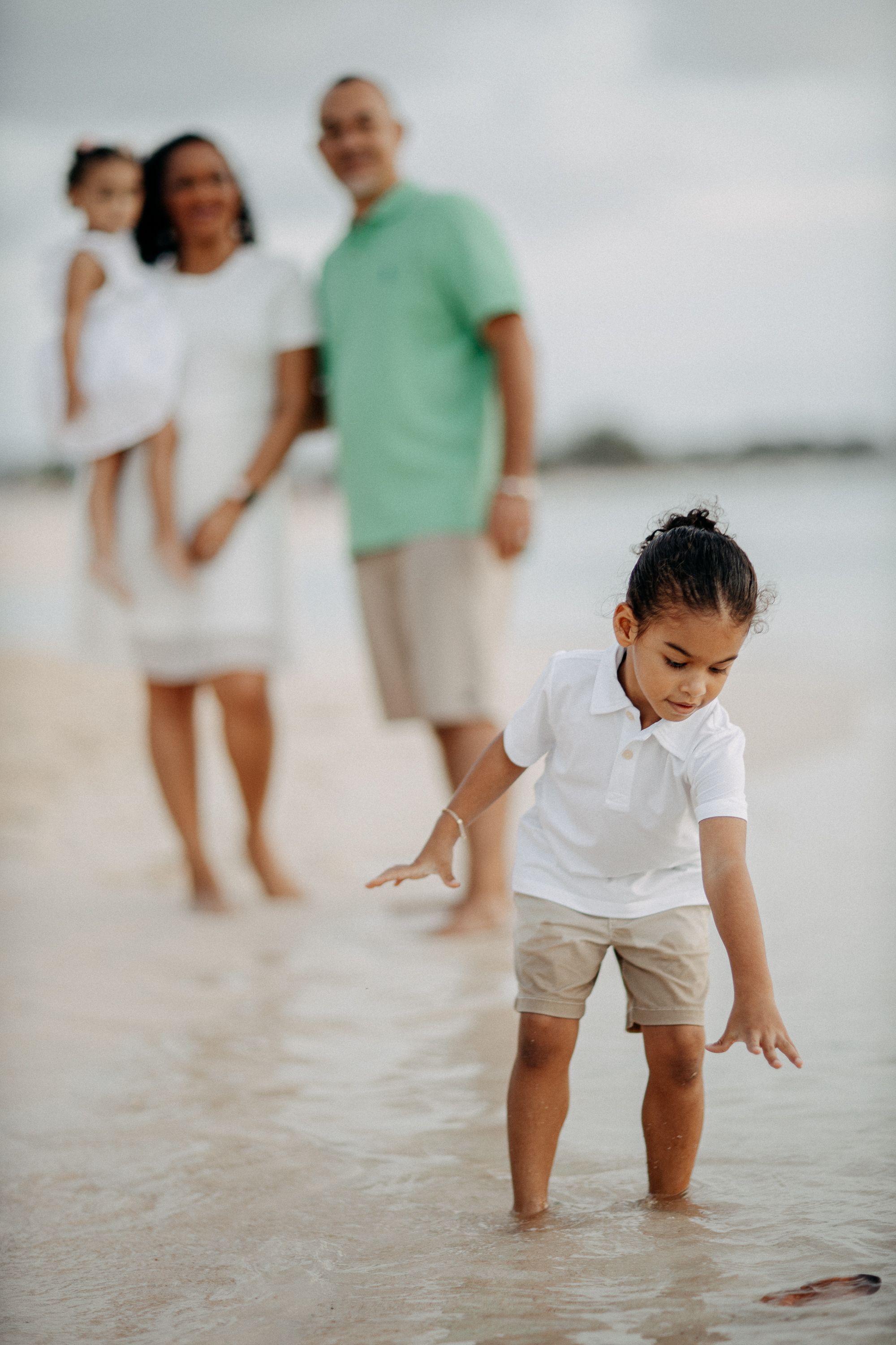 A child playing in shallow ocean water while others watch from behind on the beach.