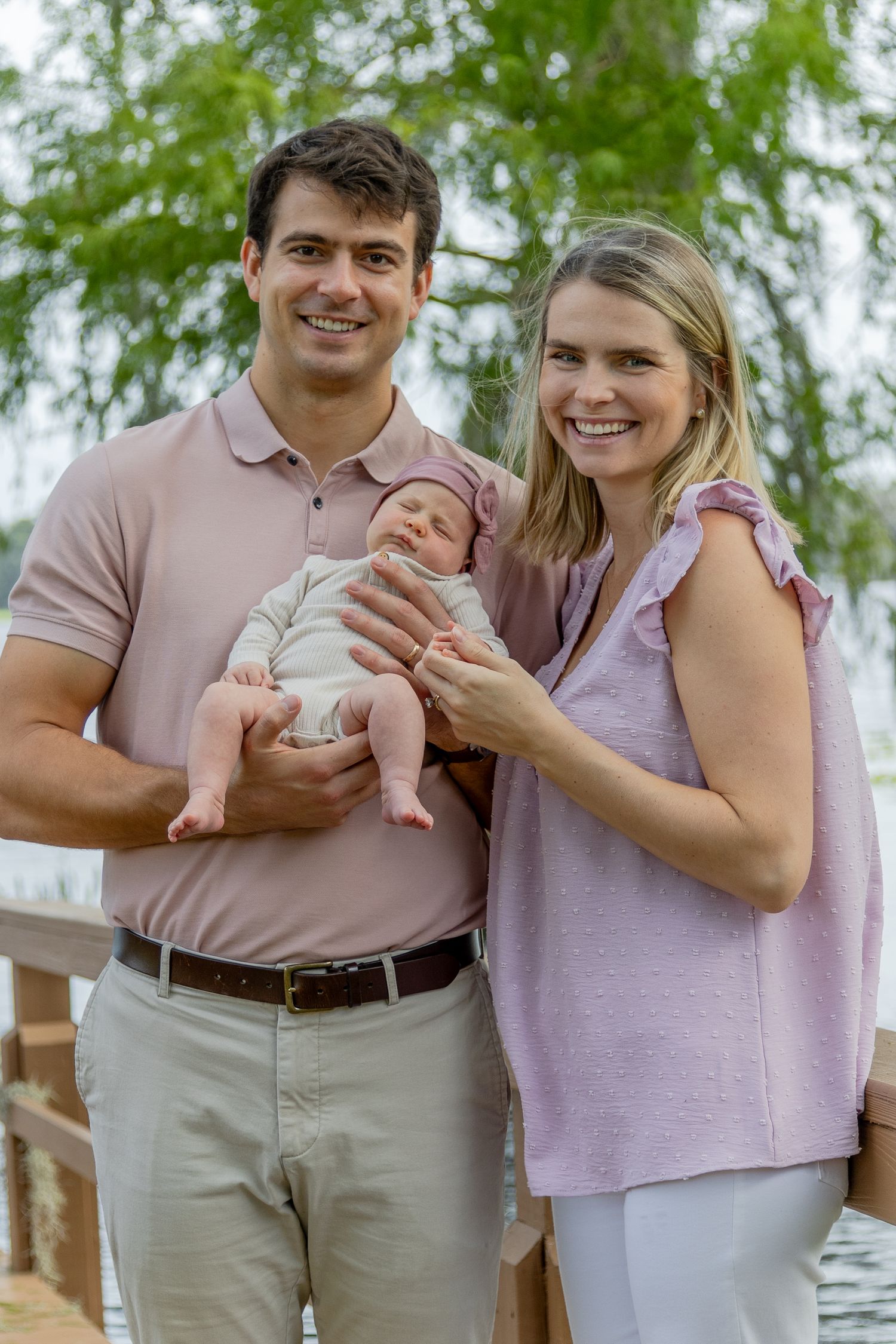 Smiling young parents holding their newborn baby on a wooden dock surrounded by trees.