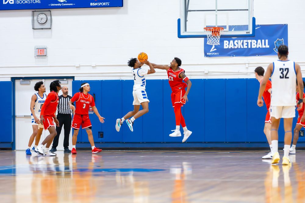Basketball players in action on a blue-trimmed indoor court with spectators watching from the sidelines.