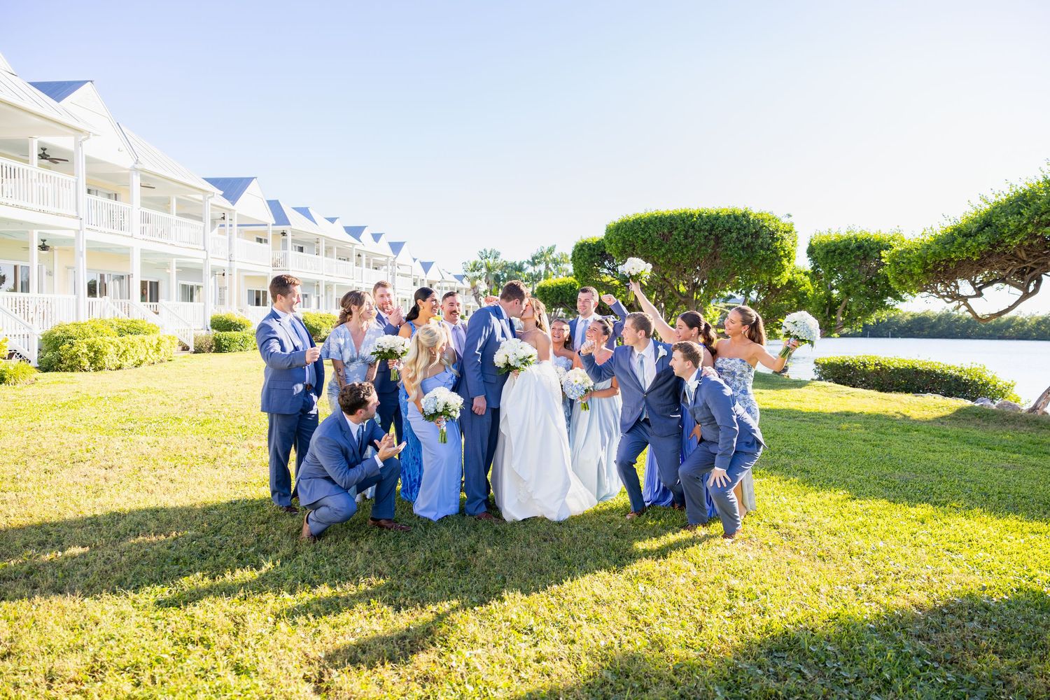 Couple shares a kiss at a beach wedding ceremony with blue floral decorations and guests seated on white chairs.