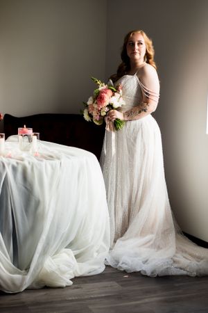 Elegant bridal portrait with flowing white dress and pink floral bouquet against grey backdrop.