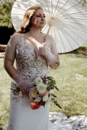 A bride holding a white parasol and floral bouquet outdoors in a floral embellished dress.