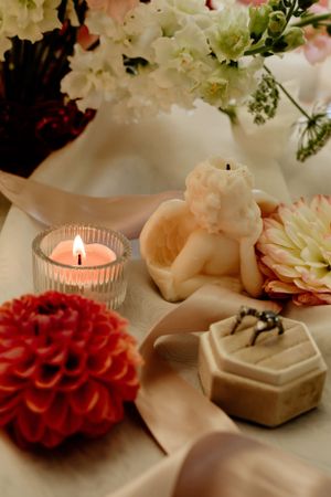 Wedding detail shot of flowers, candles and ring boxes arranged on a soft fabric surface.