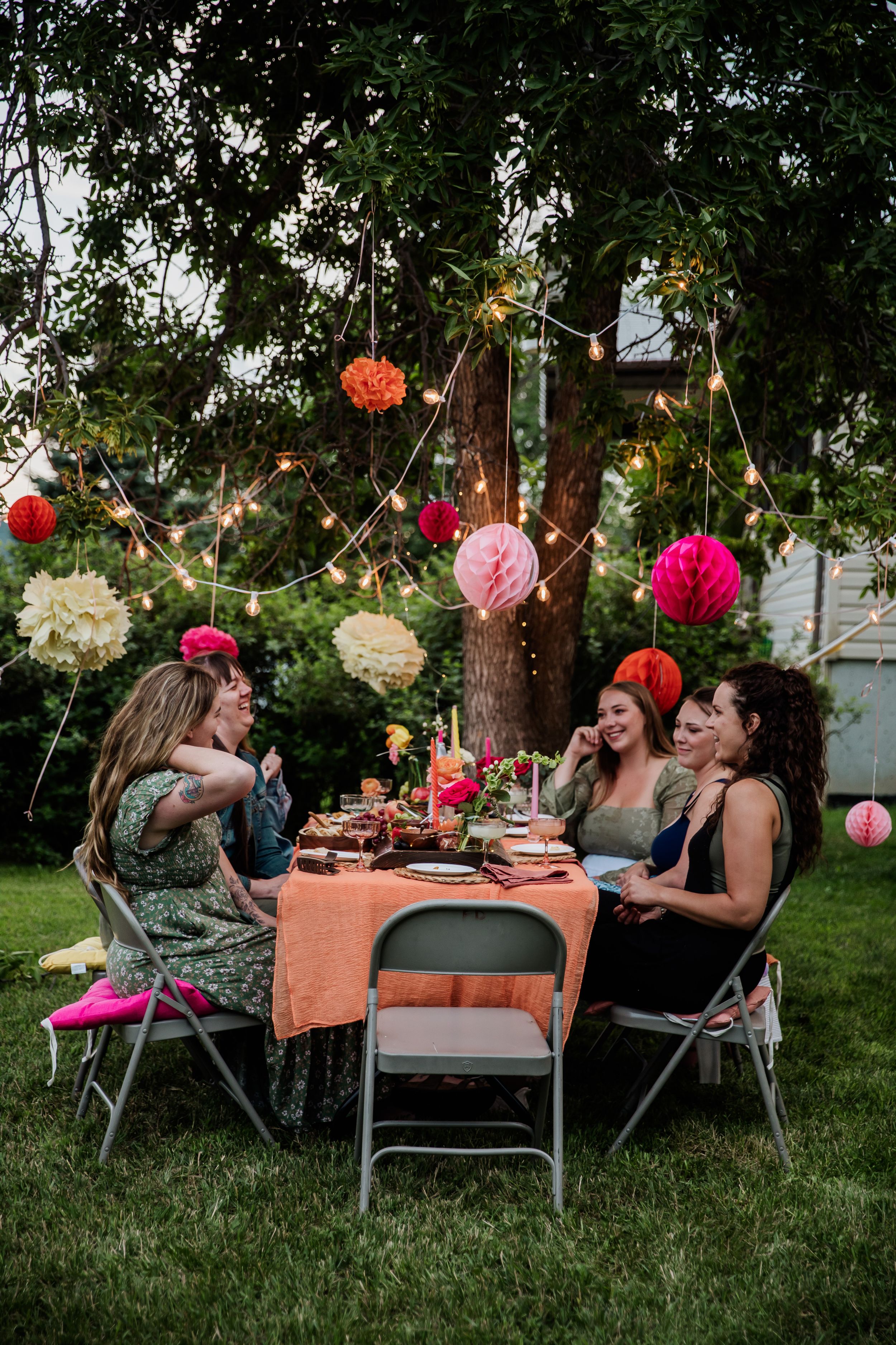 Friends gather at an outdoor dinner party under hanging paper lanterns and string lights.