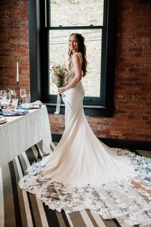 A bride in a fitted white wedding gown with a long train poses by a window against exposed brick walls.