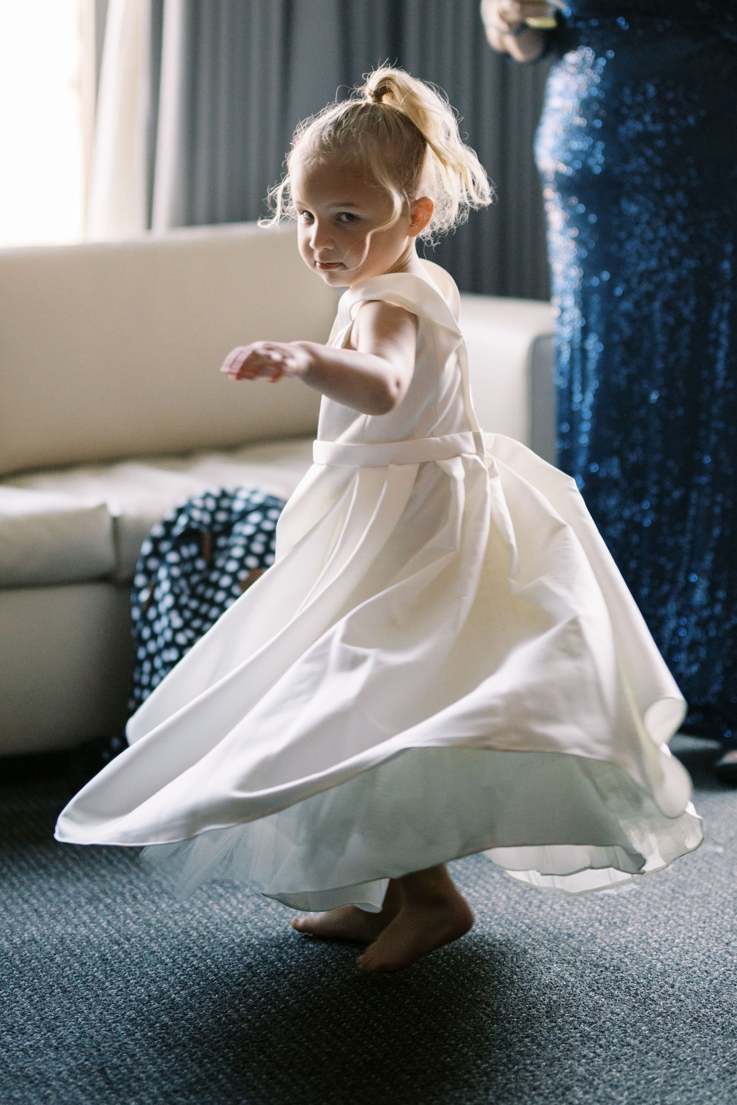 Small toddler in flowing white dress twirls and dances on carpet near blue curtains in soft window light.
