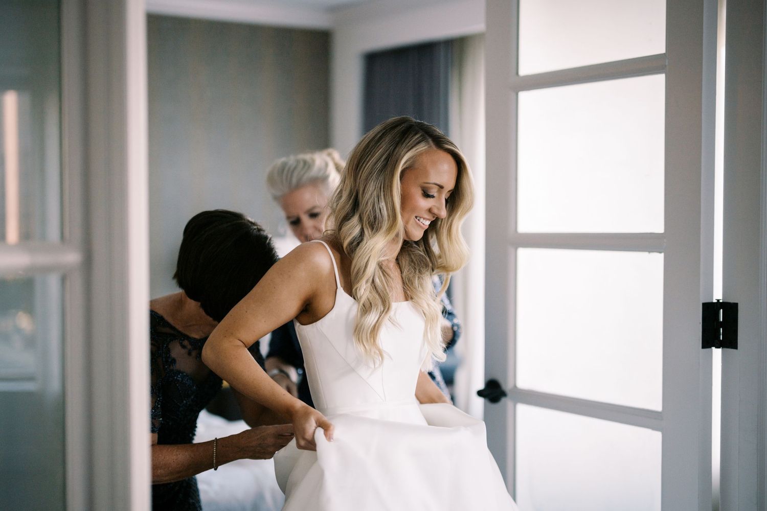 A bride gets help with her elegant white wedding dress while standing in a bright sunlit doorway.