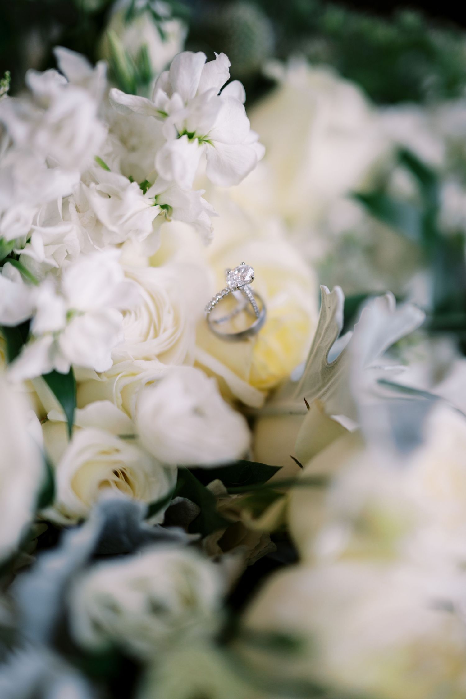 Diamond engagement ring nestled among soft white roses and flowers in a romantic wedding bouquet arrangement.
