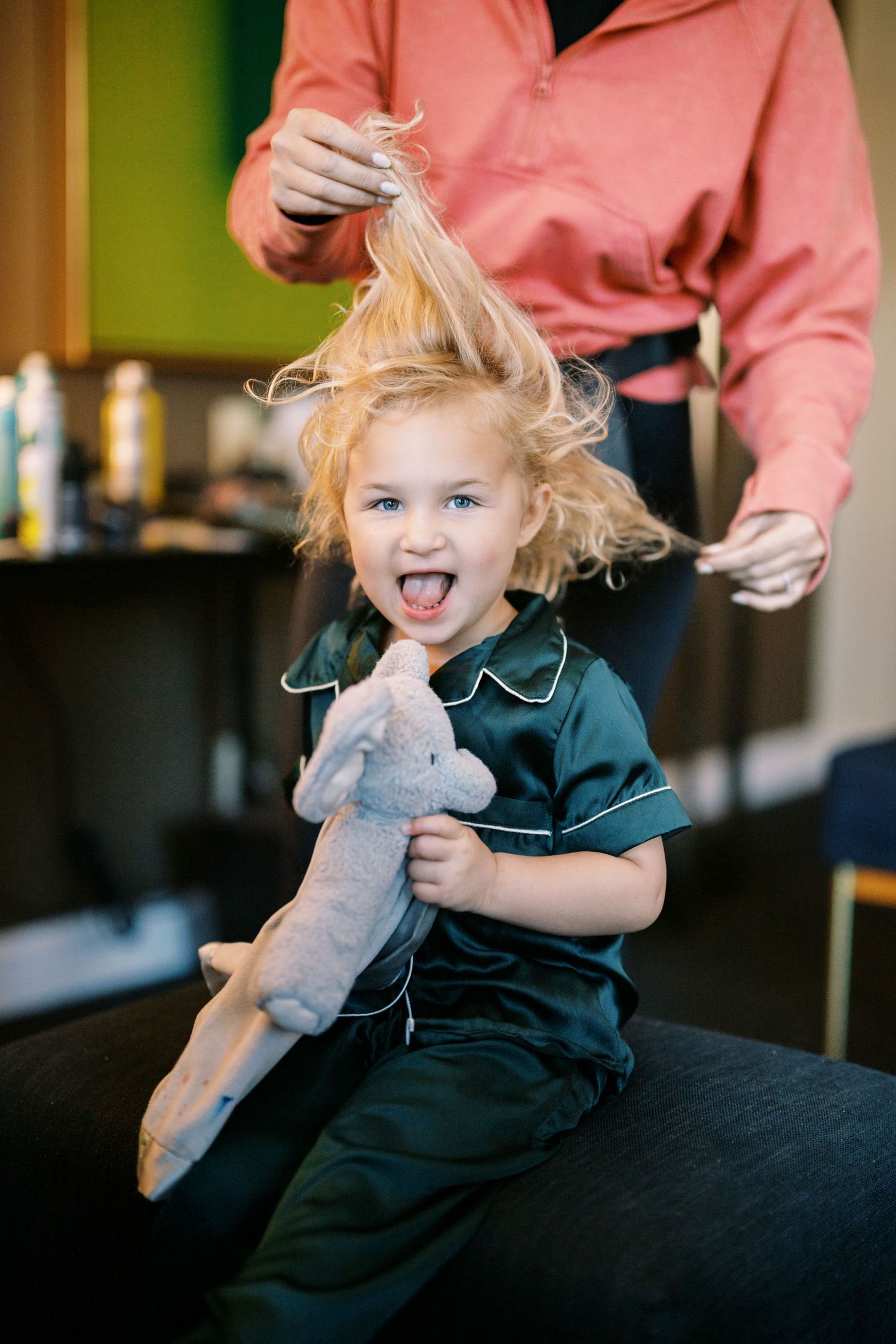 A child with messy blonde hair sticks their tongue out playfully while holding a plush toy during a haircut.