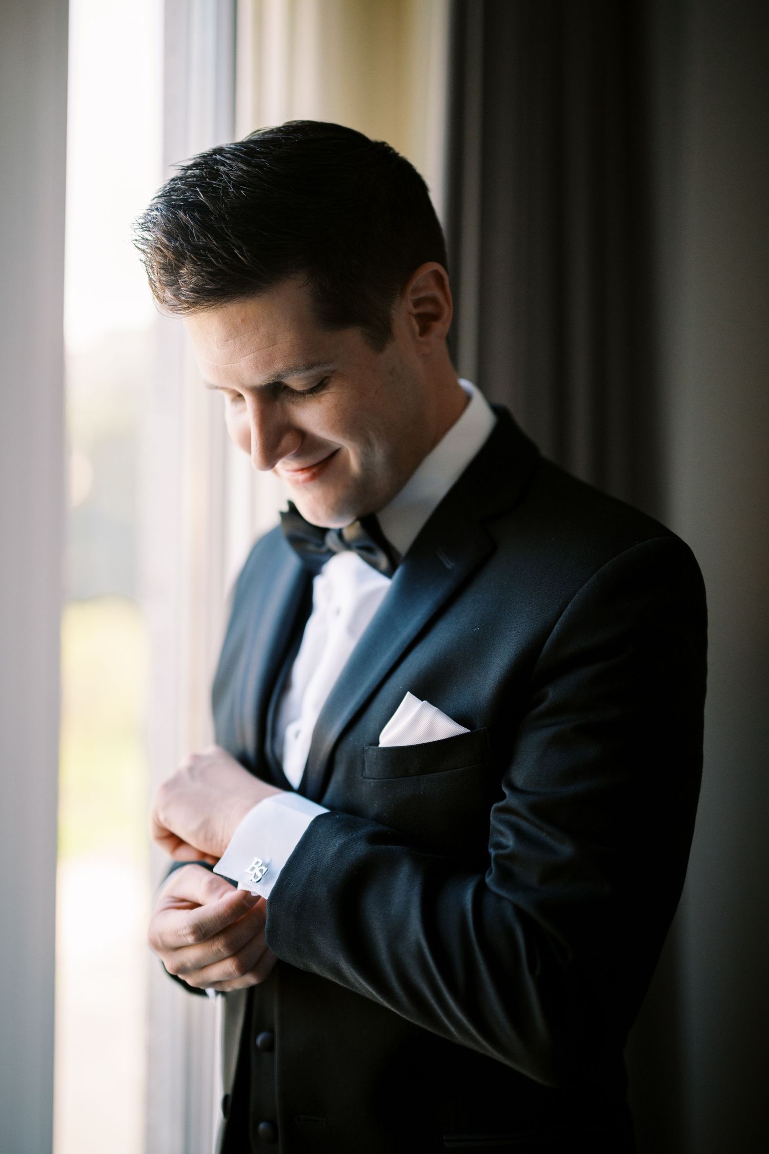 Off-center elegant profile view of a groom in a navy tuxedo adjusting his cuff by a window in soft natural light.