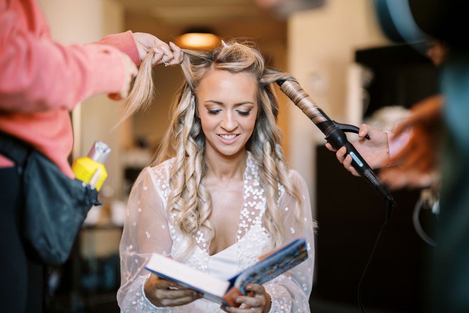 Bride reads a book while having her hair styled by two stylists during wedding preparations.