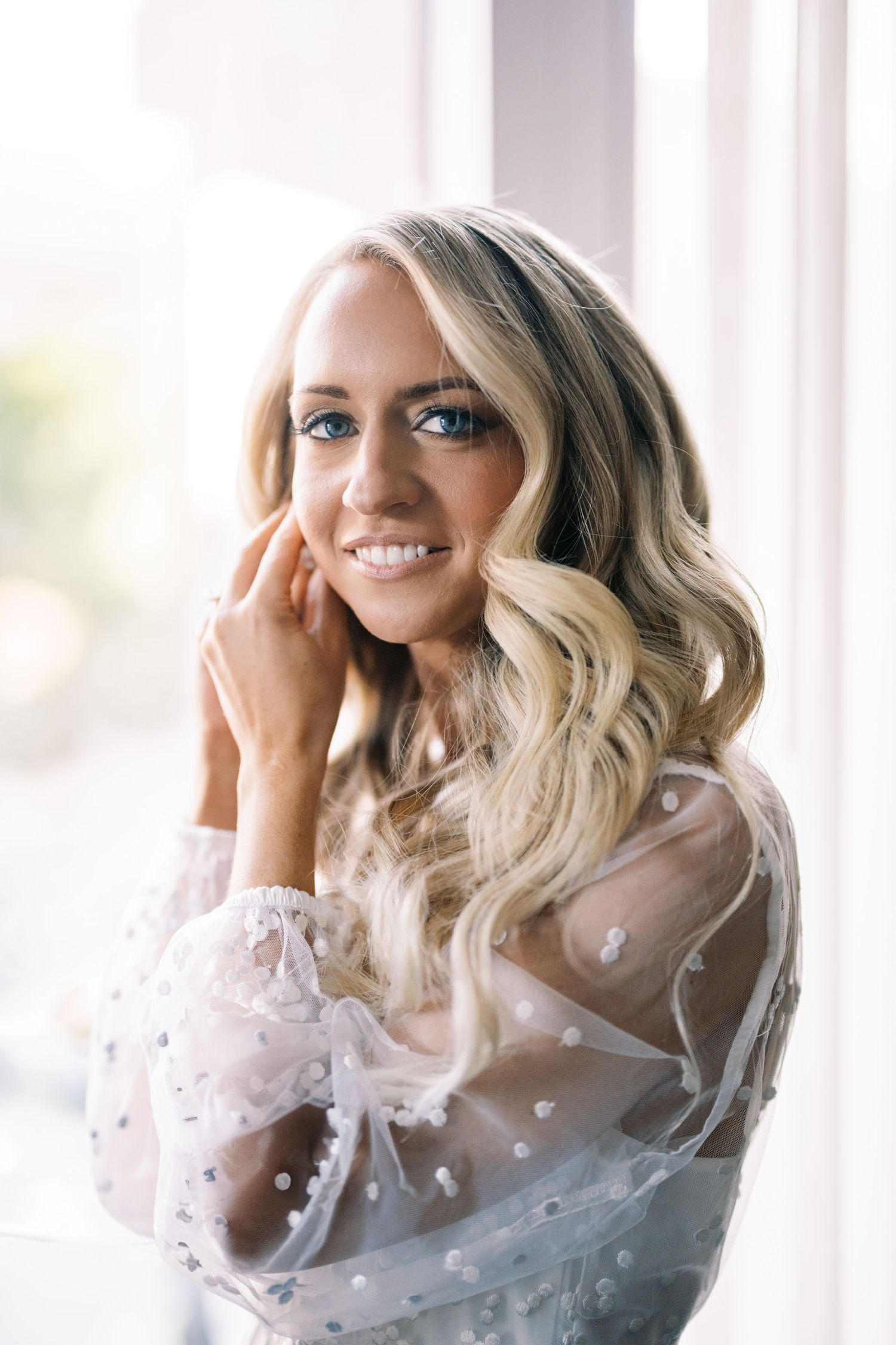 Softly lit portrait showing wavy blonde hair and sheer white polka dot top with natural window light.
