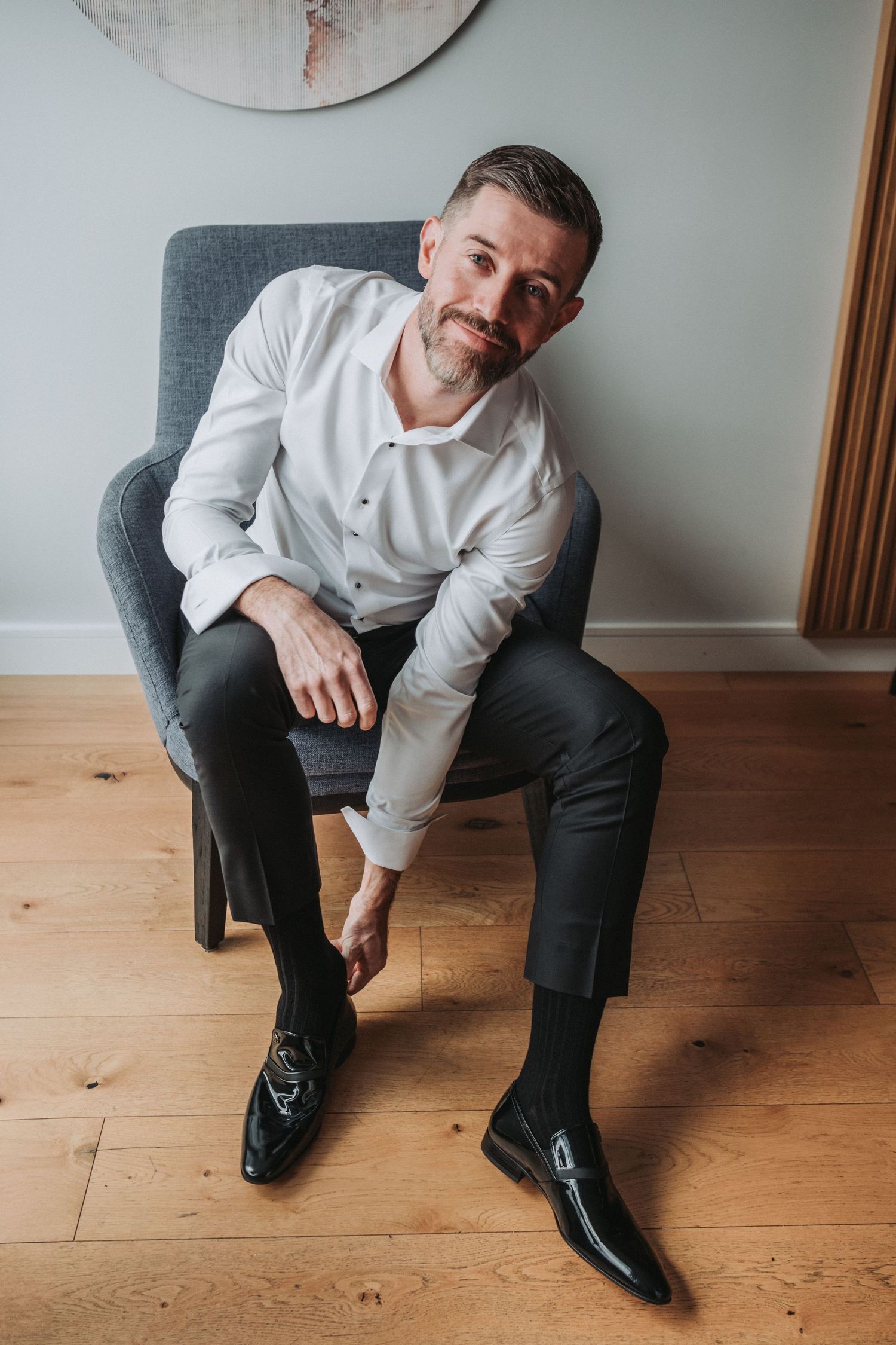 A groom-to-be is wearing a crisp white shirt and dark trousers sits casually in a modern grey armchair on hardwood flooring.