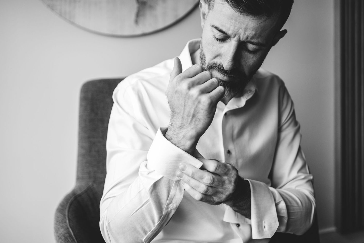 Black and white portrait of a groom-to-be in a white shirt sitting in chair and adjusting sleeve button.  Shot at the Canopy Hotel.