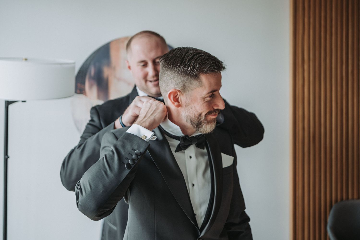 The groom-to-be gets help from his brother adjusting his bowtie and collar on a groom wearing a classic black tuxedo in a well-lit room.