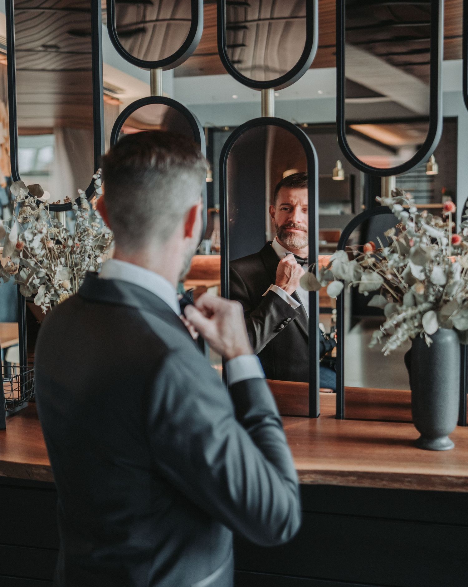 A groom-to-be in a dark suit adjusts his tie while looking in a decorative mirror in a room with floral arrangements.  Canopy Hotel.