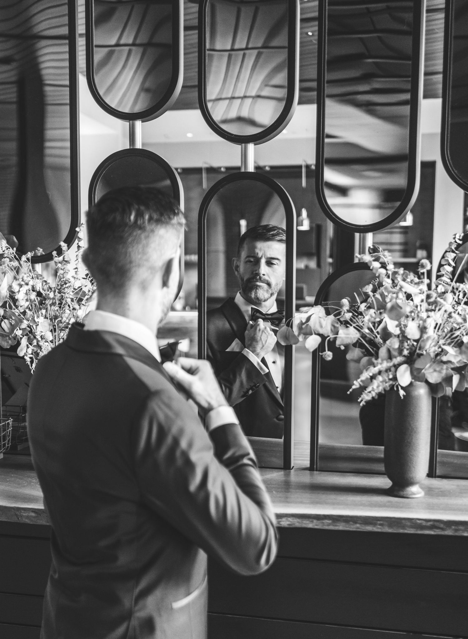A black and white photo showing a groom-to-be in formal attire looking at their reflection in an elegant mirror with floral arrangements.