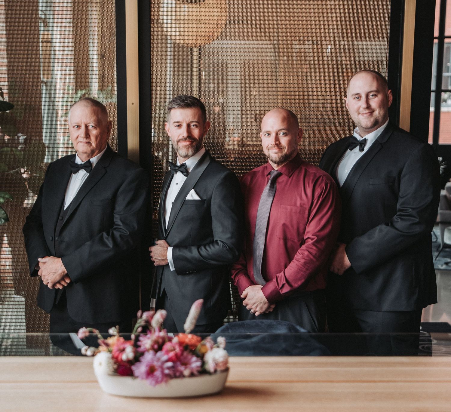 Four men in formal attire stand together at the Canopy hotel lounge in Portland, ME with a decorative floral arrangement in the foreground.