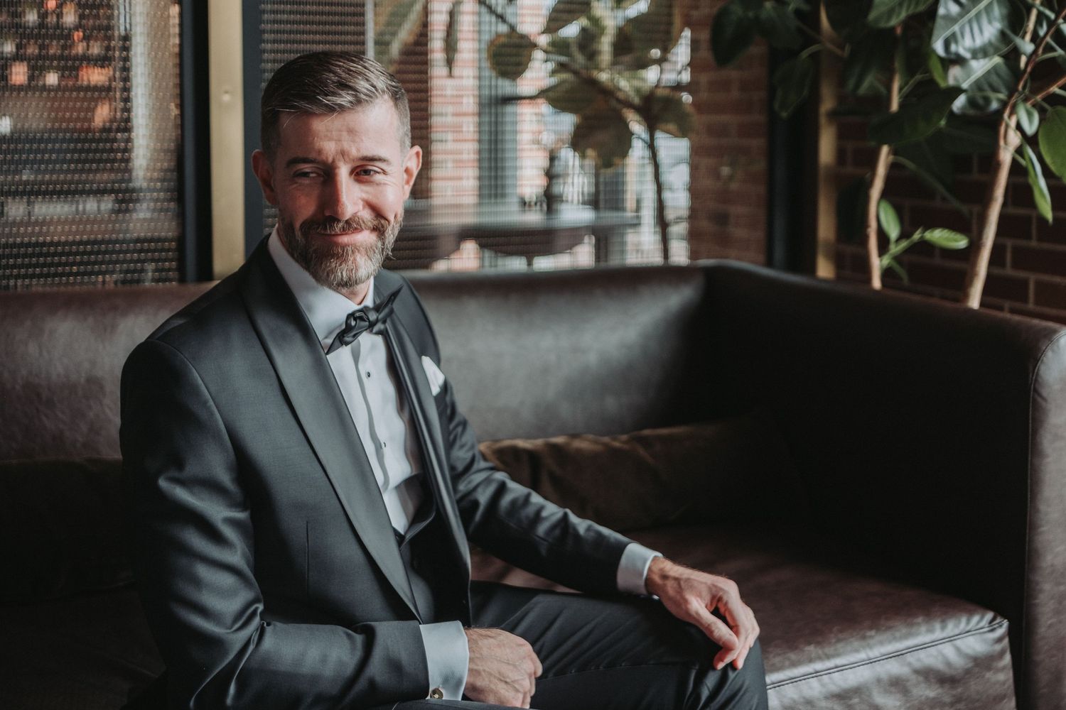 A well-dressed groom-to-be in a gray suit and bow tie sits on a leather couch in a space with exposed brick walls.  Shot at the Canopy Hotel