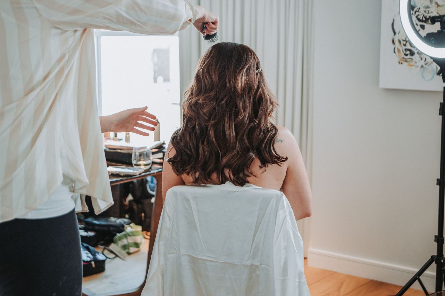 A rear view shot of a bride-to-be getting her long wavy brown hair styled in a bright white room with a mirror.  Taken at the Canopy Hotel.