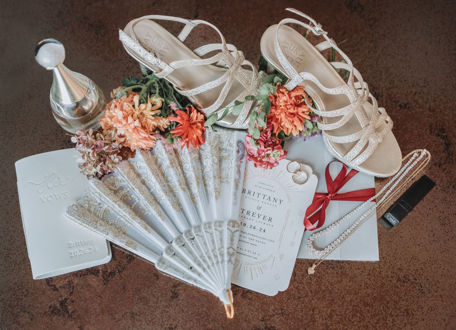 Wedding accessories including white sandals, delicate fan, floral decorations and invitation card arranged on dark backdrop.