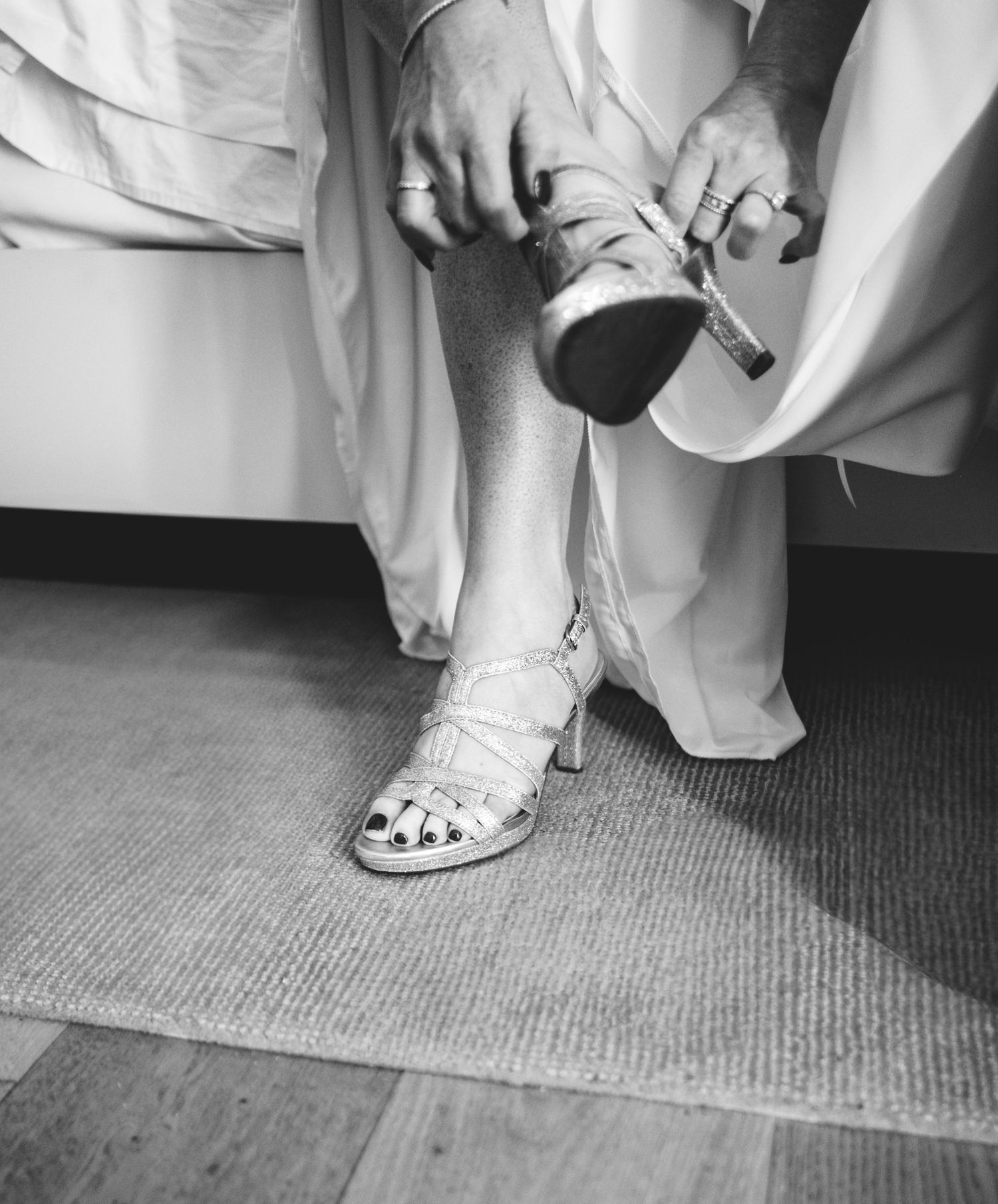A Black and white photo of a bride-to-be's hands adjusting a vintage white heeled sandal on a carpeted floor.  Located at the Canopy Hotel.
