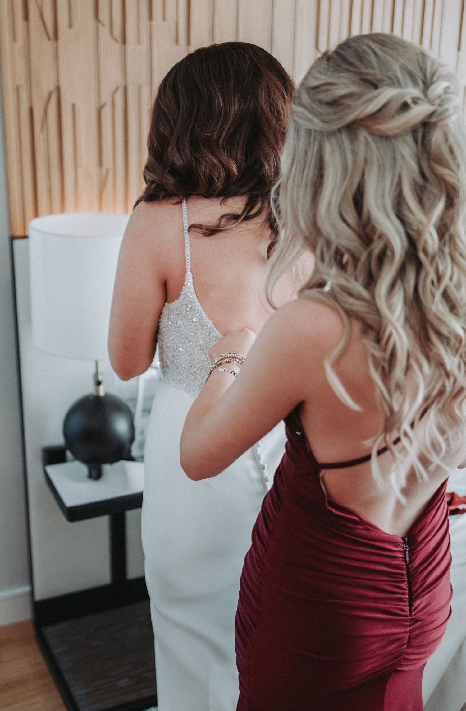 Two women in formal dresses, one in white sparkly wedding gown and one in burgundy, seen from behind in an elegant setting getting ready.