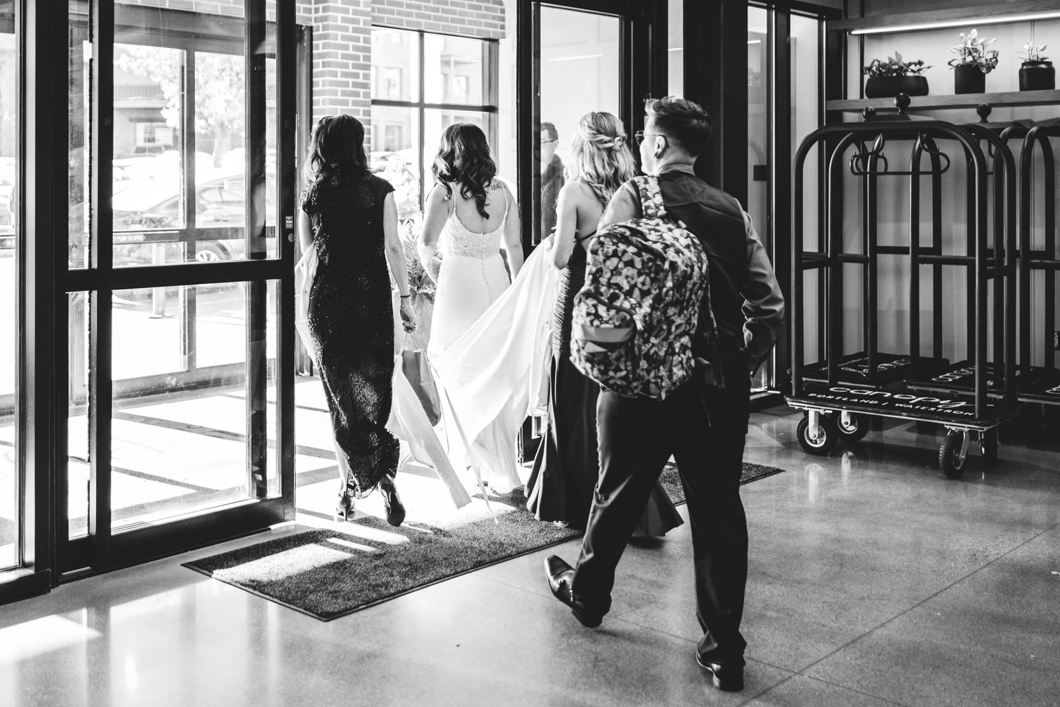 A group of friends exit the hotel lobby with luggage carts visible in black and white.  They are walking the bride-to-be to her limo.