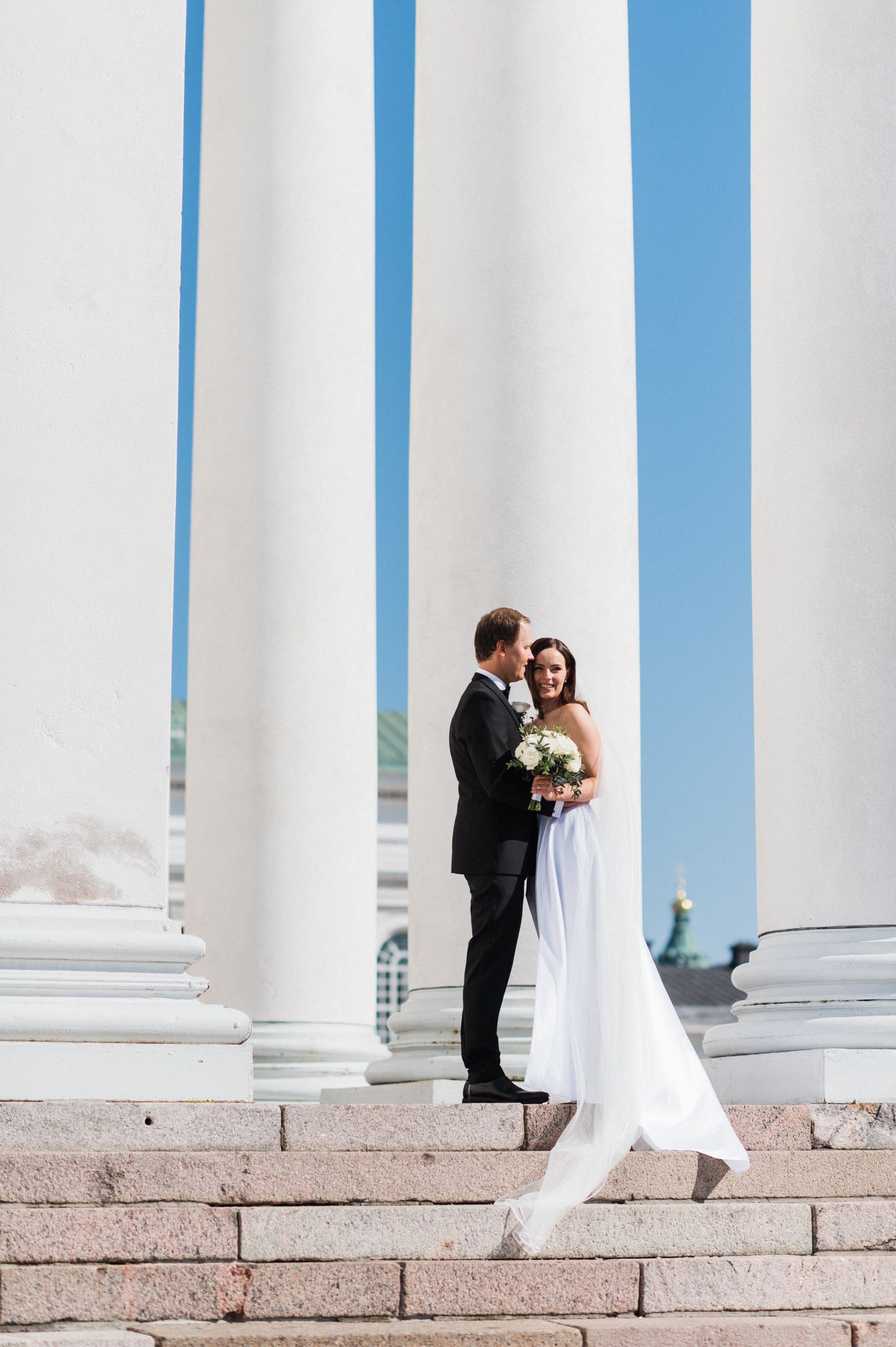 A couple shares a romantic moment between white columns against a blue sky on their wedding day.