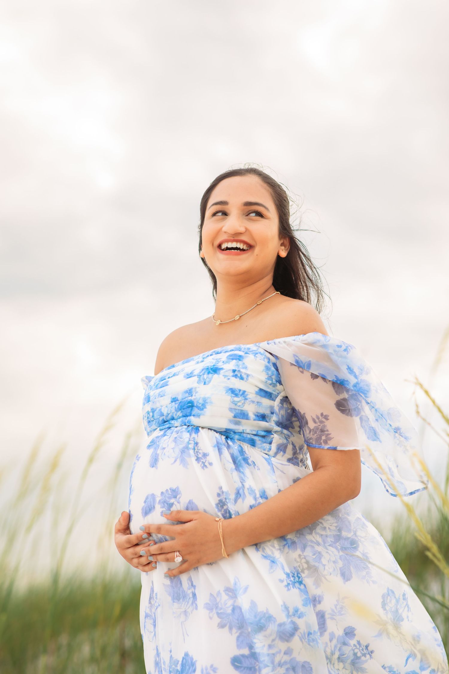 Woman wearing blue floral dress standing in coastal grass at sunset.