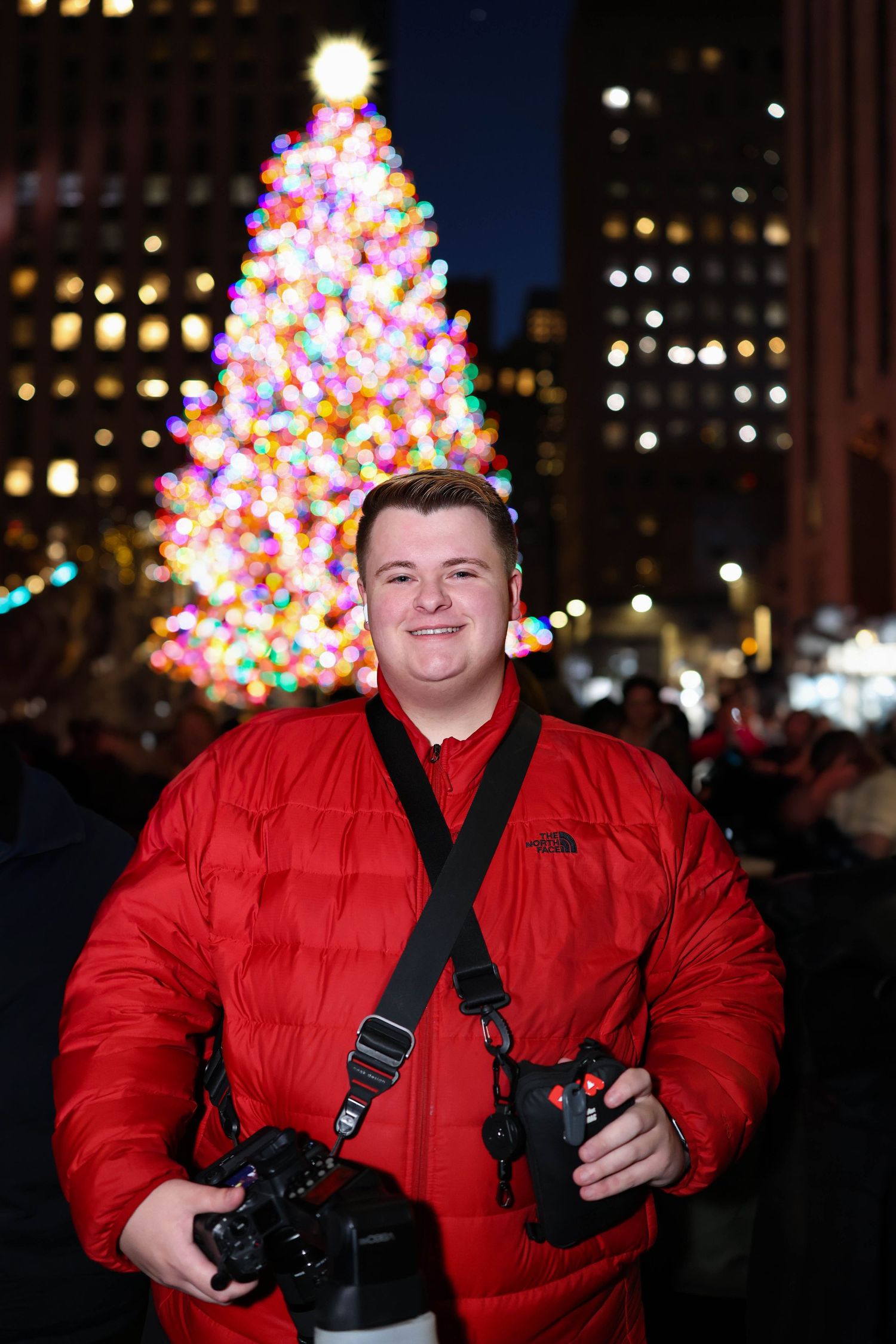 Professional photographer in a bright red winter coat holding cameras against a nighttime Christmas scene.