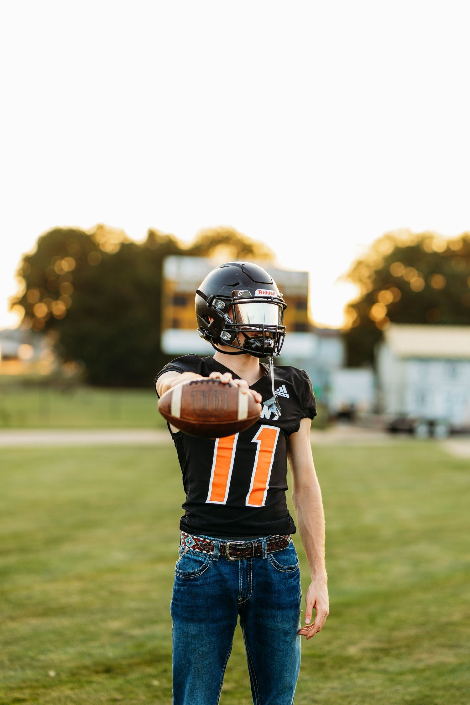 High school football player in black helmet extending ball toward camera on sunny field.