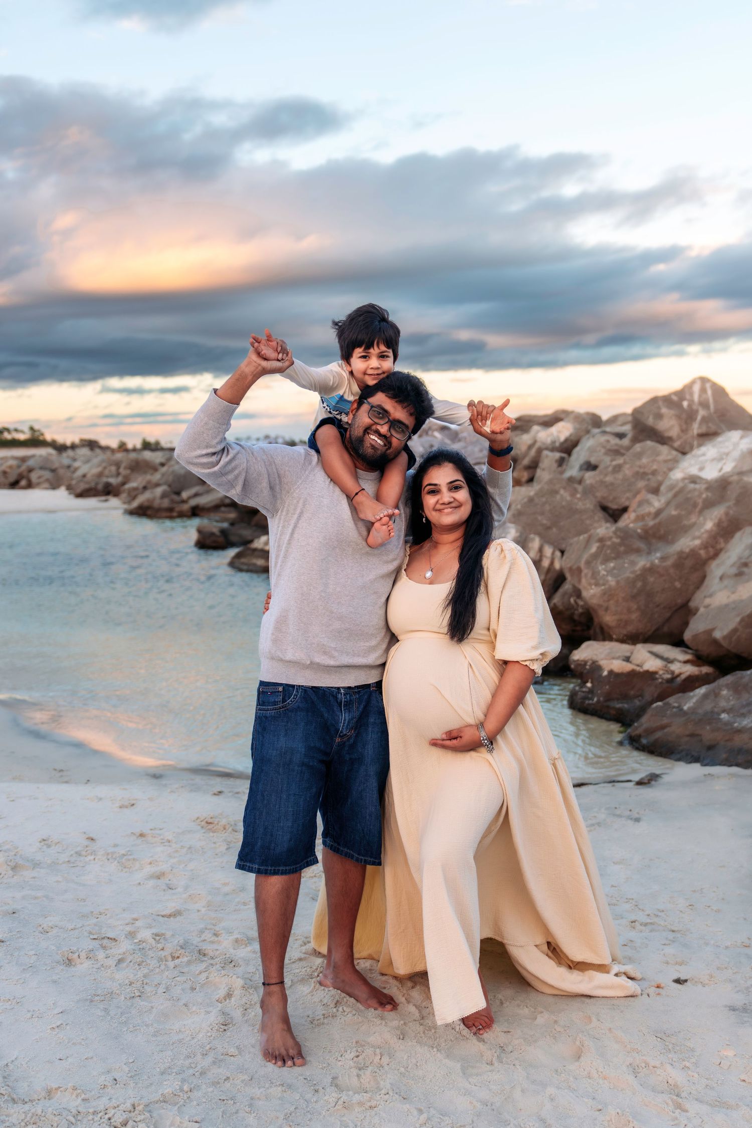 Happy family with pregnant mother in cream gown and child on fathers shoulders posing at rocky beach sunset.