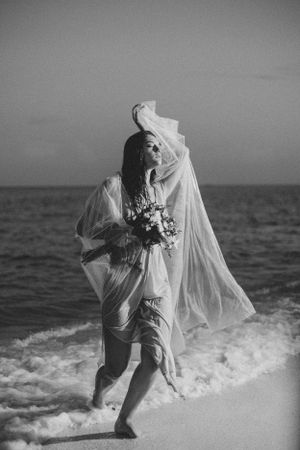 A figure in a flowing white veil dances gracefully along the ocean shore in black and white photography.