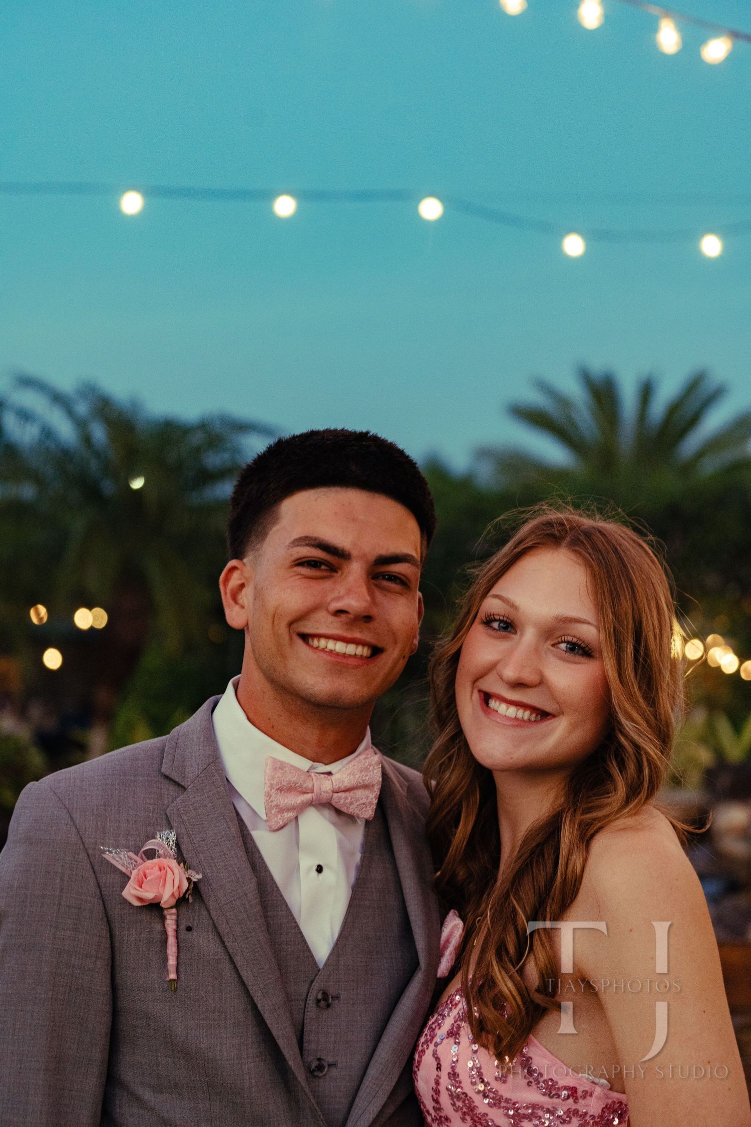 A young couple in formal prom attire smile under string lights at dusk.