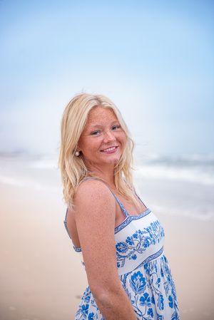 Smiling blonde woman in blue floral sundress posing on a sandy beach with ocean waves in the background.