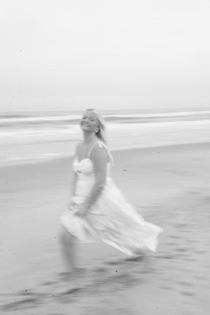 Black and white photo of a smiling woman in a flowing white dress running on a beach with ocean waves.