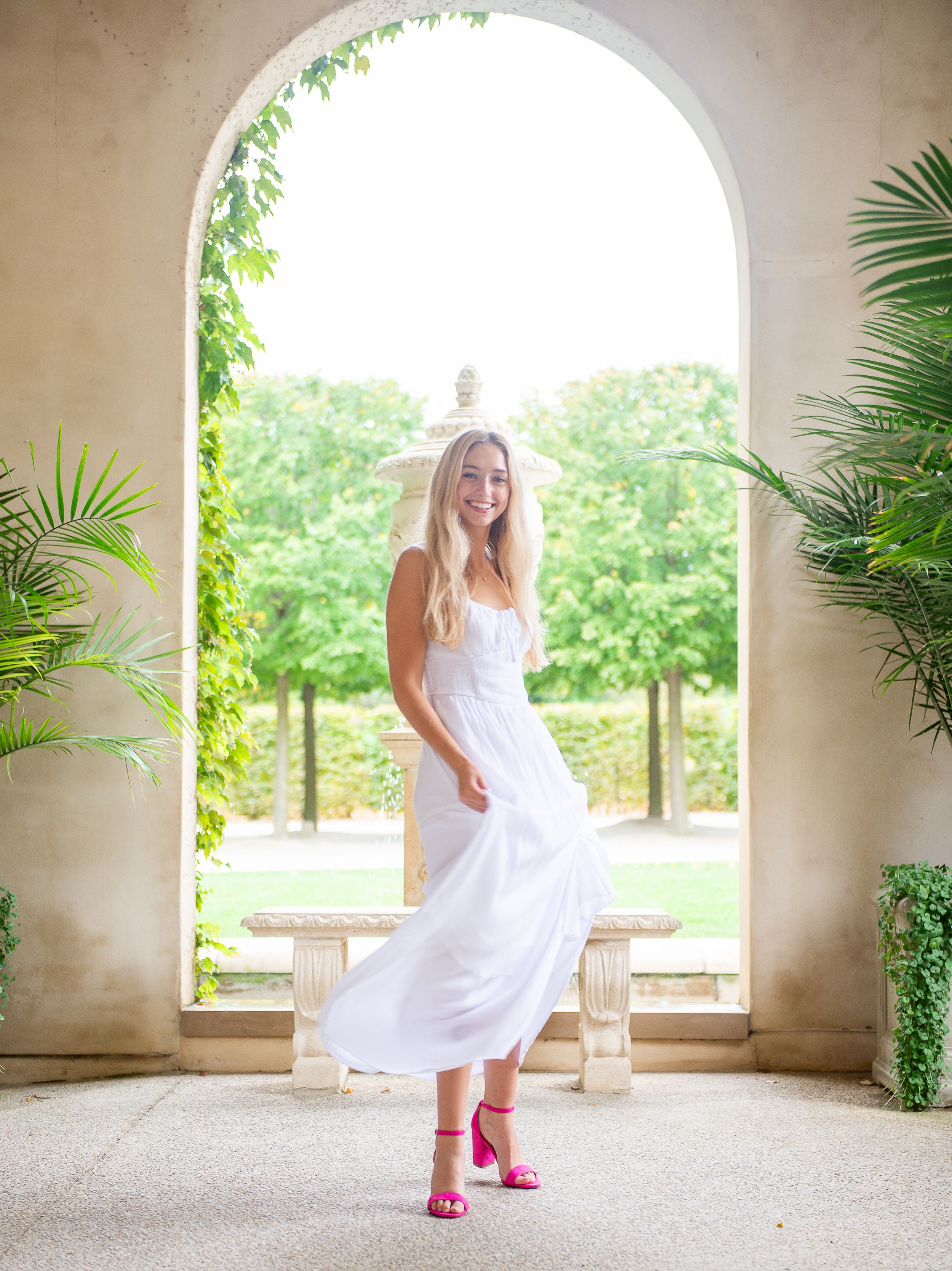 High School Senior in flowing white dress and pink shoes twirling under an arched pavilion.
