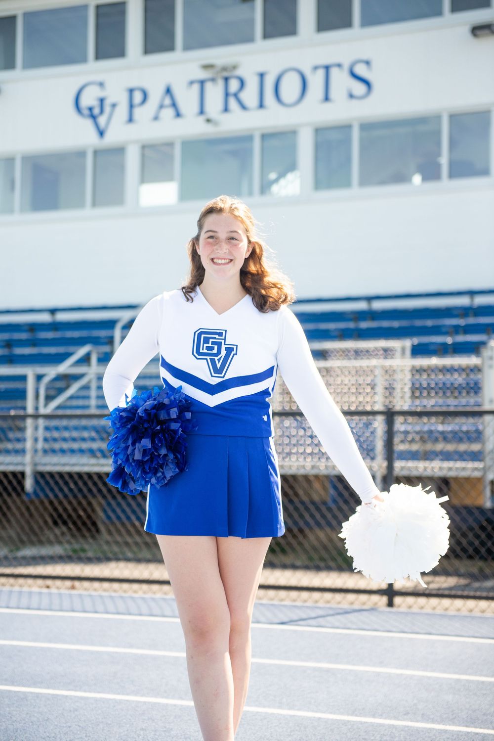 Cheerleader with pom poms posing on track field in front of Patriots stadium.