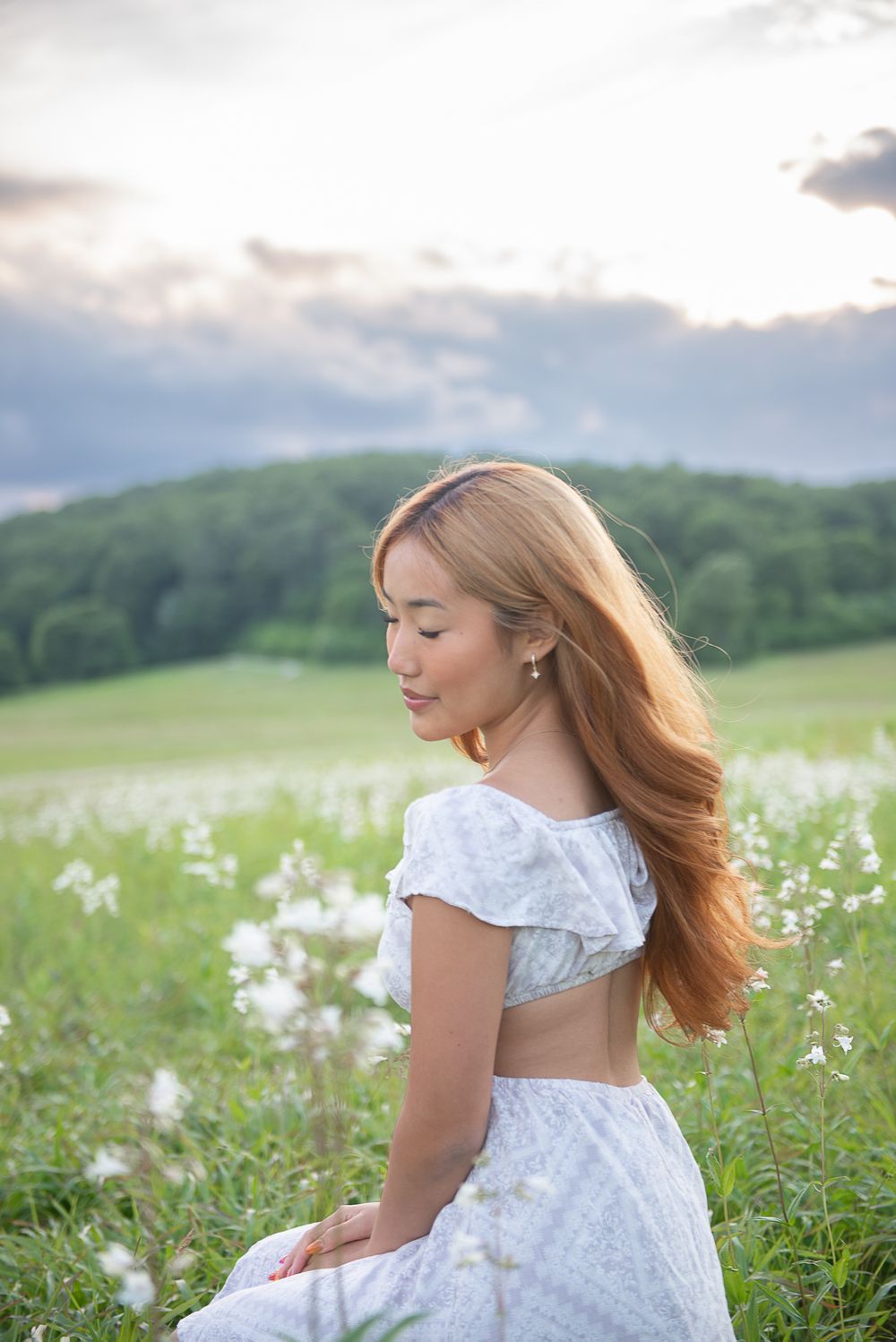 Young woman in white crop top and skirt sitting in a field of white wildflowers at sunset.