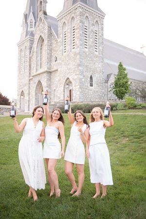 Four women in white dresses holding wine bottles outside a stone church on a sunny day.
