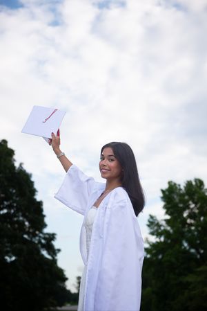 Graduate in white gown celebrating with diploma against cloudy sky.