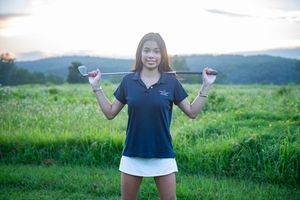 Golfer in a navy and white uniform, holding a gold club in front of sunset on a golf course.