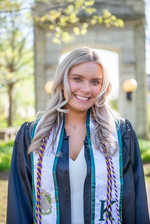 Graduate wearing ceremonial stole and gown poses outdoors with decorative columns in background.
